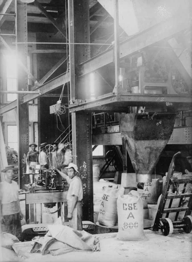 Black and white photo of Filipino working inside a factory, surrounded by steel beams and sacks of sugar.