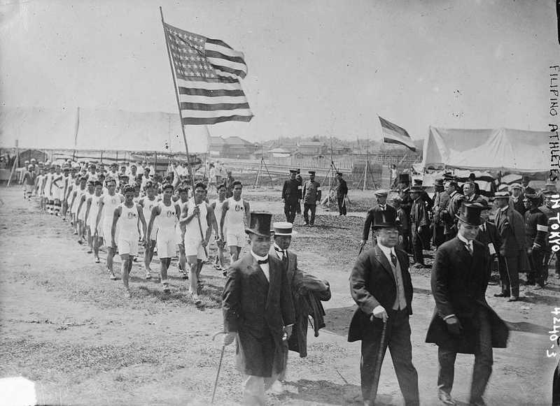 Black and white photo of Americans in suits marching through field followed by Filipinos in white folding an American flag.