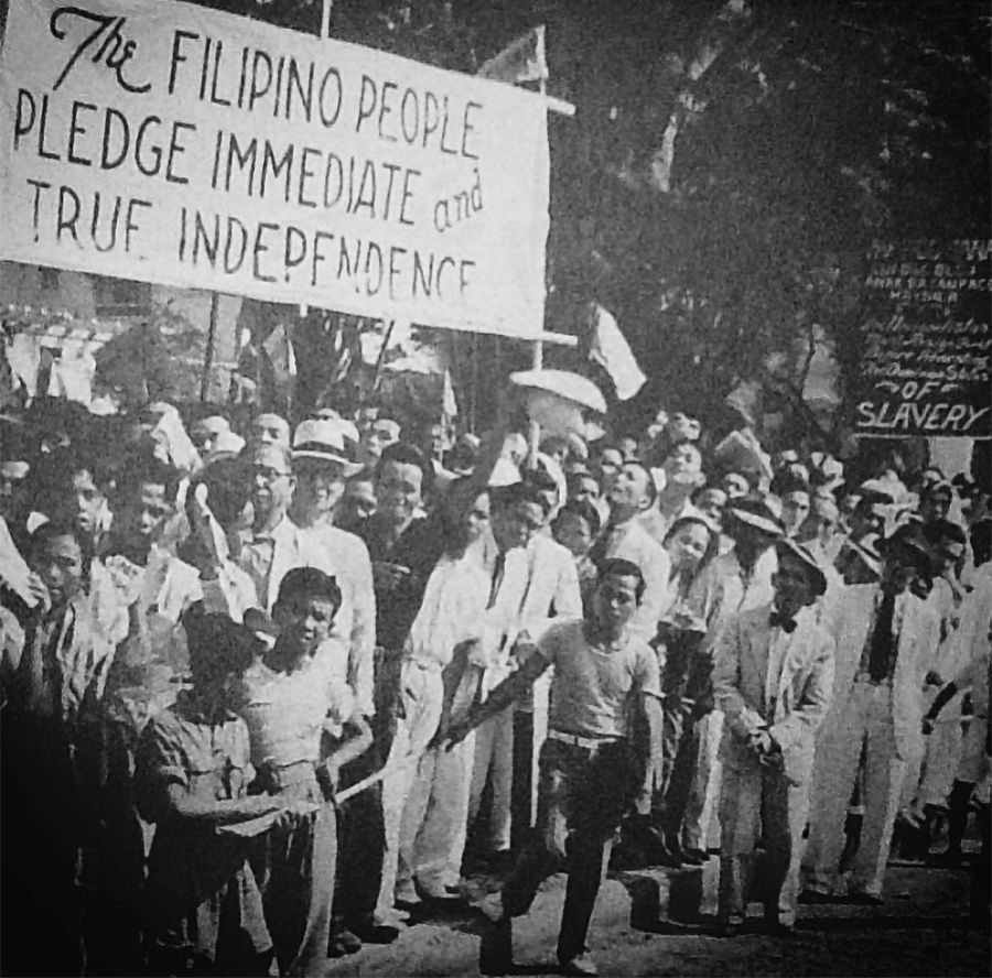 Black and white photo of crowd of protesting Filipinos holding up signs, including one that reads, 