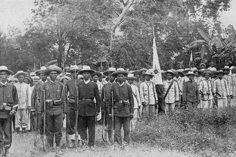 Black and white photo of Filipino revolutionaries in uniform, carrying rifles and flag
