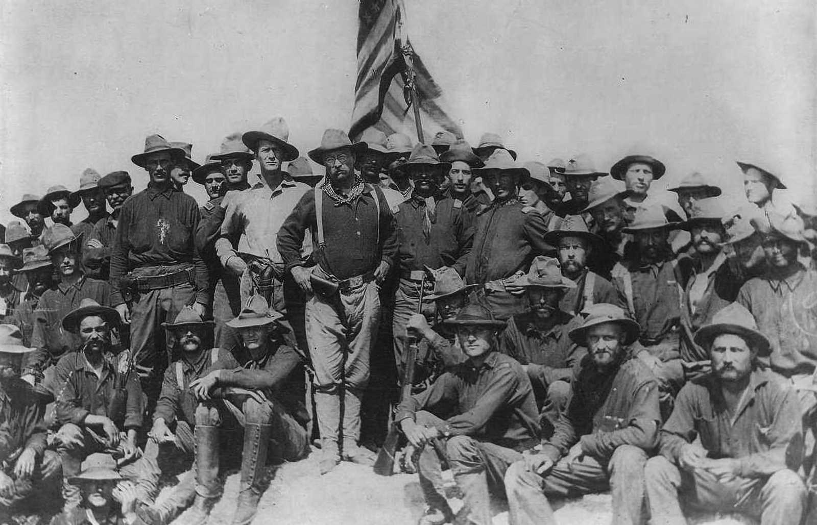 Black and white photo of Theodore Roosevelt surrounded by troops sitting on a hill, with the American flag hoisted behind them