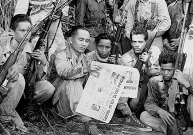 Image of Luis Taruc holding a newspaper while surrounded by other soldiers overlooking and holding rifles