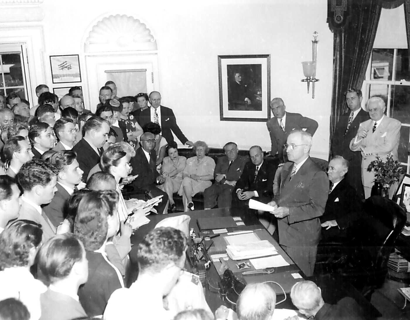 Photo of Harry Truman standing behind desk with other officials facing crowd of reporters
