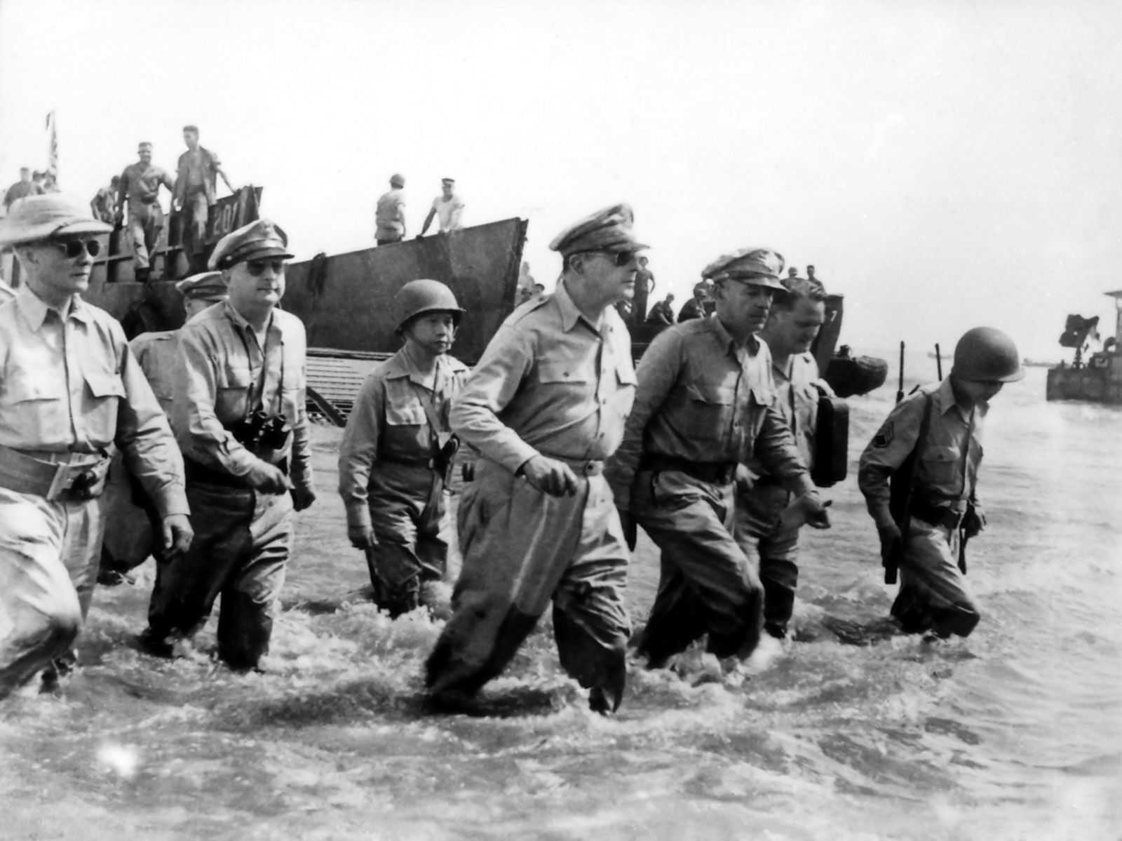 Photo of Douglas MacArthur surrounded by officers wading through water on a beach with ships and soldiers behind