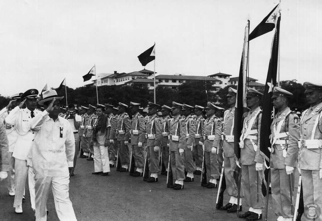 Photo of white-suited officers standing and waving at soldiers who stand at attention and raise the Philippine flag