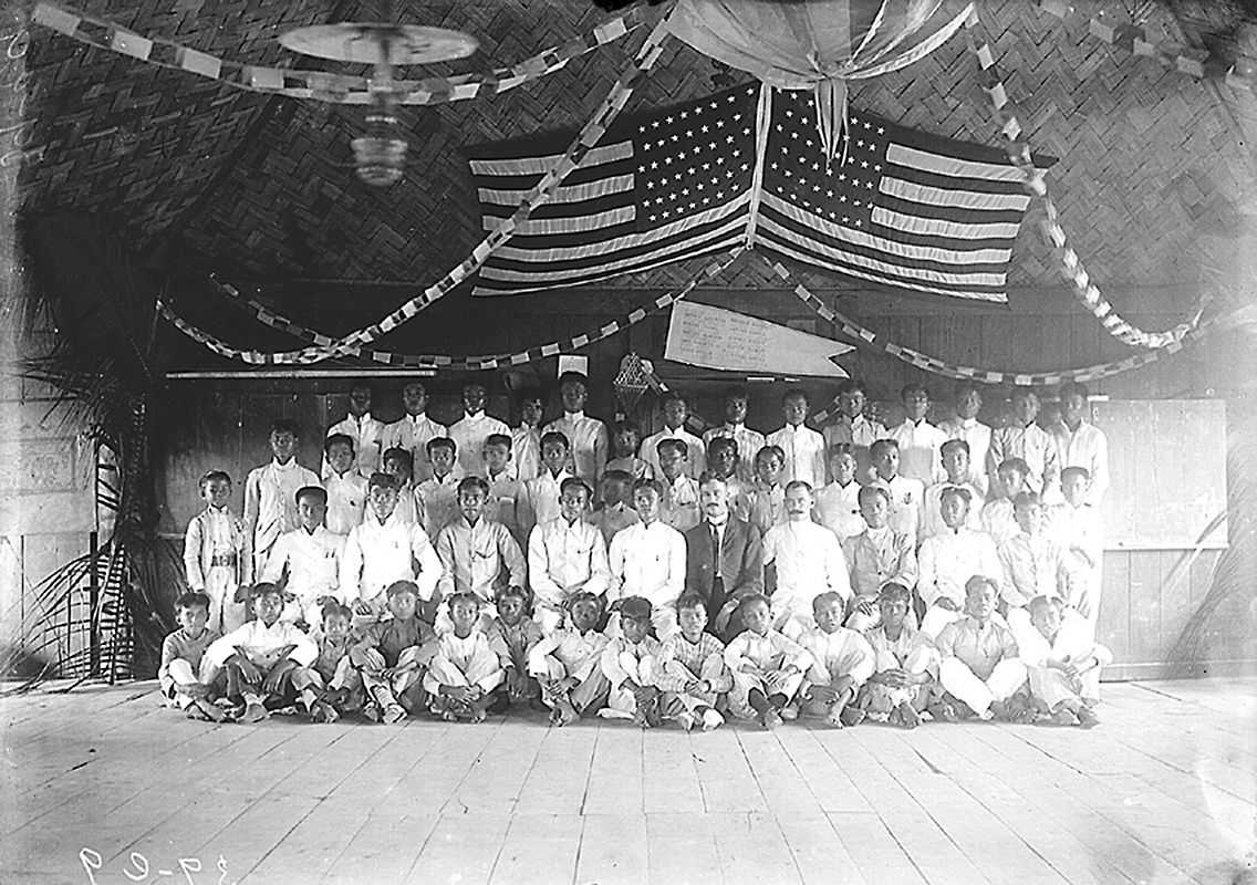 Black and white photo of Filipino students posing for class photo with American teacher and American flags above them