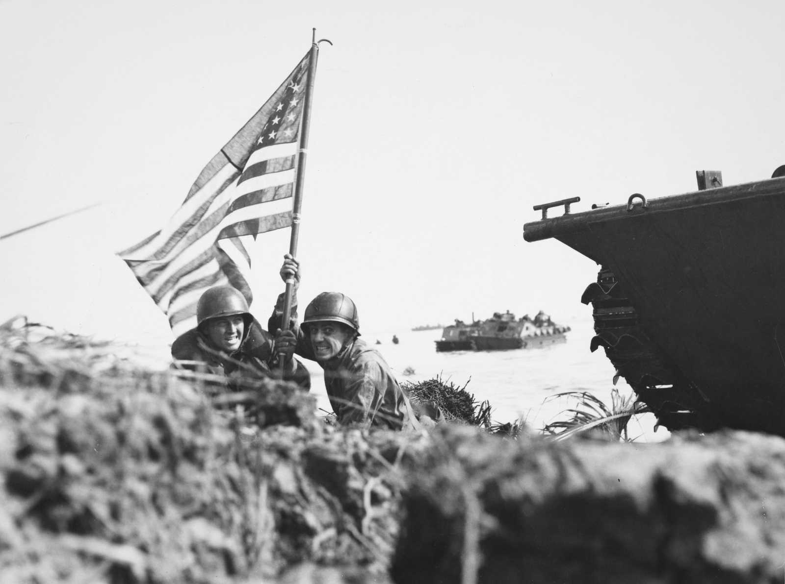Photo of two soldiers landing on a beach raising an American flag, with water and ships in the background