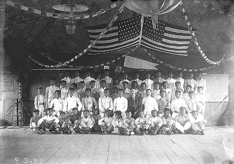 Black and white photo of Filipino students posing for class photo with American teacher and American flags above them