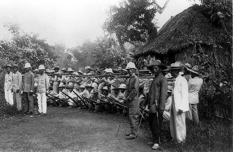 Black and white photo of Filipino revolutionary soldiers gathered in a field standing outside of a hut