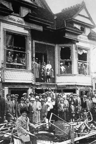 Black and white photo of Filipino Americans gathered around a house that has its windows blown out and wooden scraps littered around.