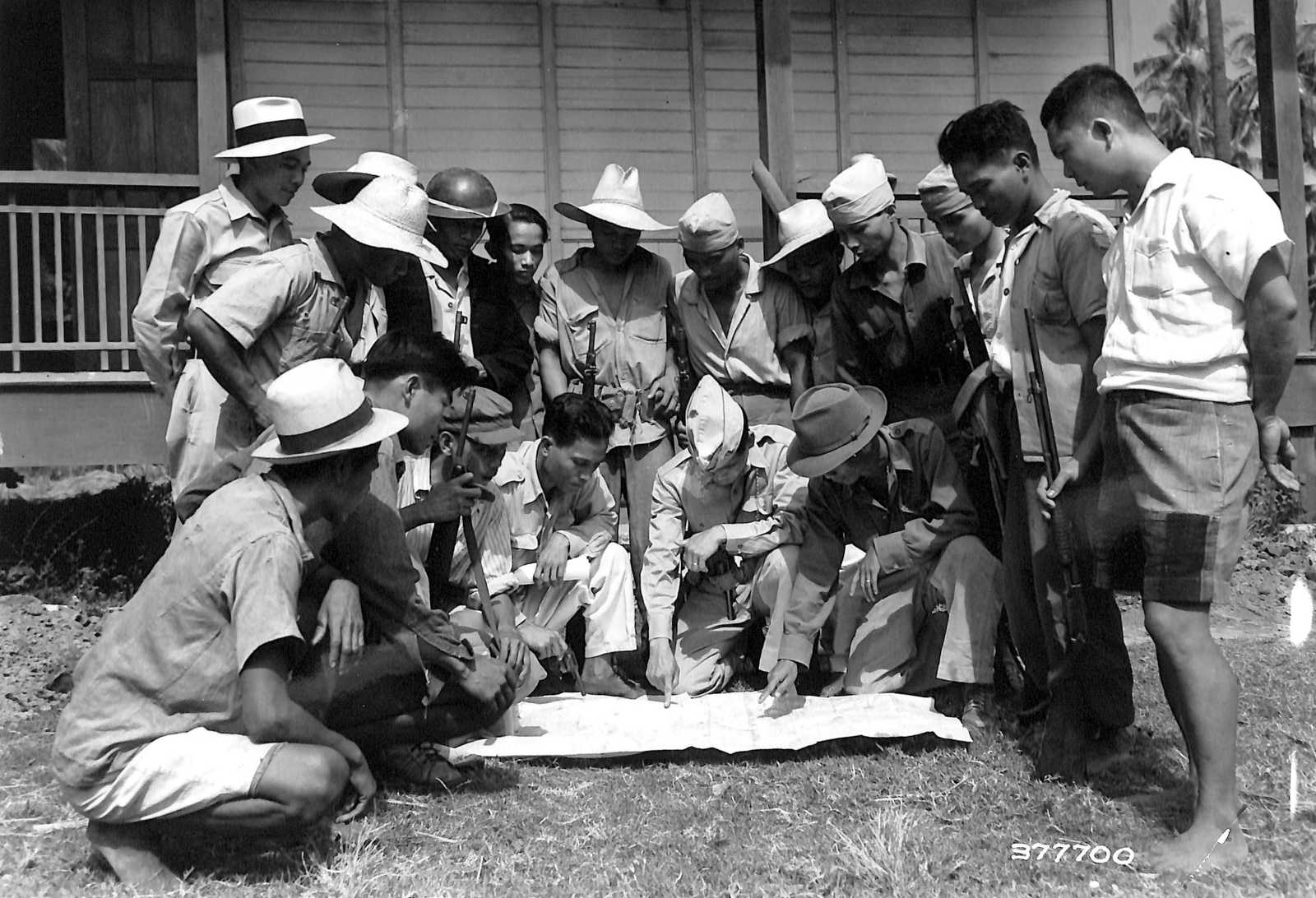 Photo of Filipino guerrillas in casual dress standing over a document being reviewed by kneeling American officers